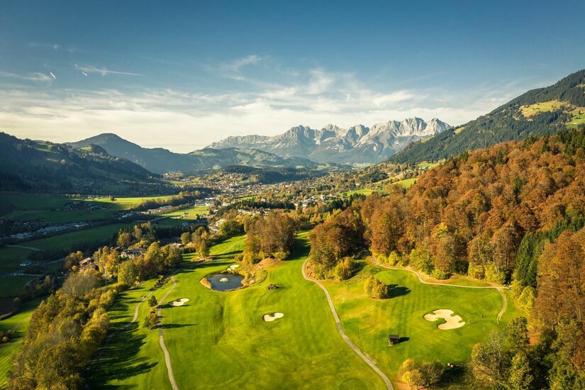 Luftaufnahme eines Golfplatzes mit herbstlichen Bäumen vor einem Bergmassiv.