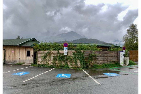 Ein Parkplatz mit behindertengerechten Stellplätzen und einem Holzgebäude im Hintergrund. Der Himmel ist bewölkt, und im Hintergrund sind Berge sichtbar.