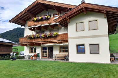 Ein traditionelles Alpenhaus mit Holzbalustraden und blühenden Blumenkästen. Im Hintergrund liegen grüne Wiesen und bewaldete Berge.