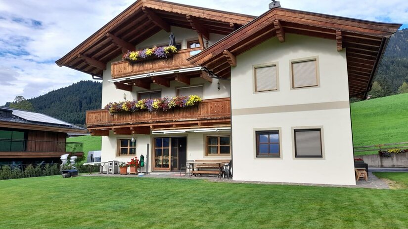 Ein traditionelles Alpenhaus mit Holzbalustraden und blühenden Blumenkästen. Im Hintergrund liegen grüne Wiesen und bewaldete Berge.