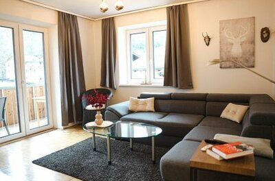 Ein modernes Wohnzimmer mit einer grauen Couch und einem Glastisch. Die großen Fenster lassen viel Licht herein und zeigen eine gemütliche Atmosphäre.