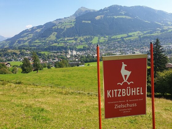 Infotafel Zielschuss Kitzbühel Rote Hinweistafel Zielschuss Kitzbühel auf einer grünen Wiese mit Blick auf den Ort und Berge.