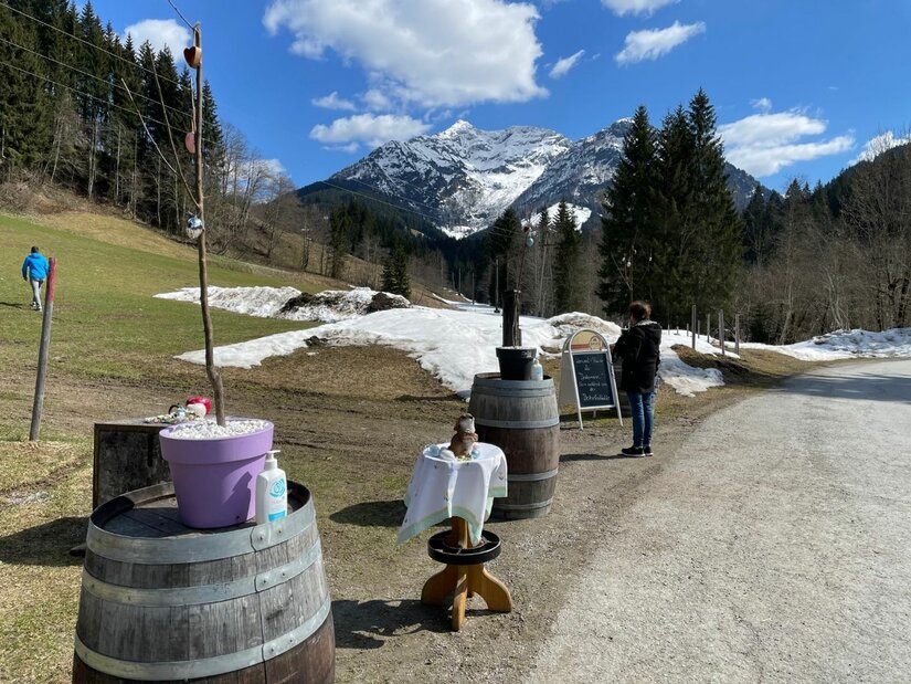 Wanderweg am Hörndlingergraben Wanderweg mit Holzfässern und einer Person vor einem schneebedeckten Berggipfel unter blauem Himmel.