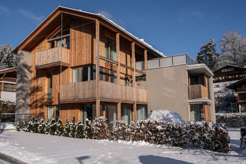 Kitzbühel Apartments Haus Wegener Modernes mehrstöckiges Holzgebäude mit Balkonen im Schnee unter blauem Himmel.