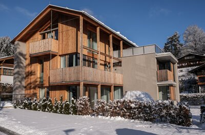 Kitzbühel Apartments Haus Wegener Modernes mehrstöckiges Holzgebäude mit Balkonen im Schnee unter blauem Himmel.