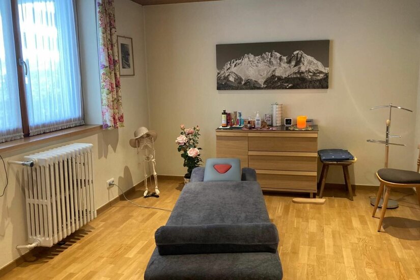 Ein Behandlungsraum mit Massageliege, Holzkommode und einem Gebirgsbild an der Wand.