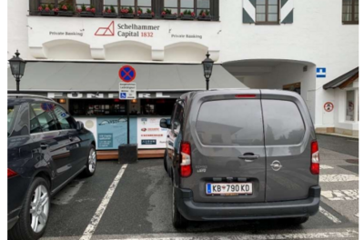 Eine graue Lieferwagen parkt vor einem Geschäft mit einem Schild „Schelhammers Capital 1832“. Im Hintergrund sind weitere Autos und ein Gebäude mit Fenstern zu sehen.
