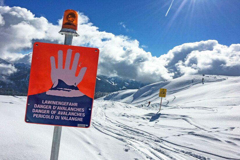 Ein Warnschild vor Lawinengefahr in einem verschneiten Berggebiet. Die Landschaft ist von Schnee und hohen Bergen umgeben, mit einem klaren Himmel im Hintergrund.