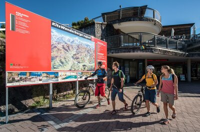 Eine Gruppe von Radfahrern steht vor einer Informationswand, die Wander- und Radwege zeigt. Im Hintergrund sind moderne Gebäude und strahlend blauer Himmel zu sehen.