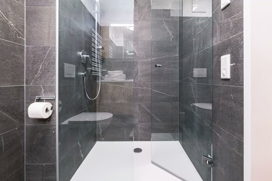 Eine moderne Dusche mit einer Glasabtrennung und grauen Fliesen. Es gibt eine Regendusche und einen Handtuchhalter.