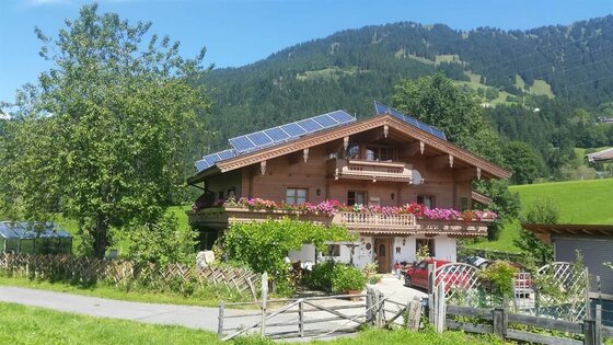 Zweistöckiges Holzhaus mit Blumenschmuck am Balkon, Photovoltaikanlage auf dem Dach und Bergen im Hintergrund.