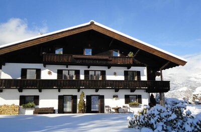 Zweistöckiges Landhaus mit Holzbalkonen und weißer Fassade in verschneiter Landschaft unter blauem Himmel.