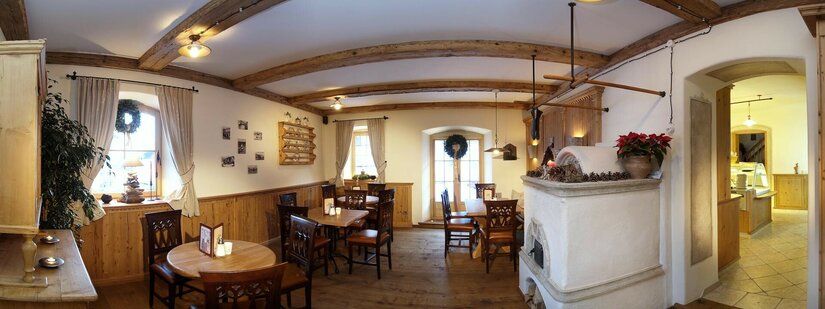 Innenansicht eines Cafés mit Holztischen, dunklen Stühlen, einem Kachelofen und Deckenbalken aus Holz.
