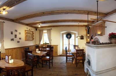 Innenansicht eines Cafés mit Holztischen, dunklen Stühlen, einem Kachelofen und Deckenbalken aus Holz.