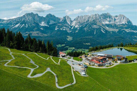 Eine malerische Landschaft mit grünen Wiesen und majestätischen Bergen im Hintergrund. Im Vordergrund liegt eine kleine Siedlung mit einem Teich und gewundenen Wegen.