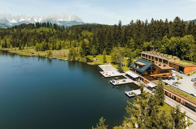 Alpenhotel Strandbad am Schwarzsee Luftaufnahme eines Holzgebäudes mit Badestegen an einem See vor Wald und Gebirgskette.