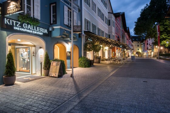 Gepflasterte Fußgängerzone in Kitzbühel mit beleuchteten Schaufenstern und historischen Fassaden in der Dämmerung.