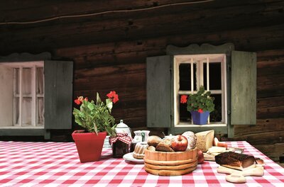 <p>Ein Tisch mit rot-weißen Tischdecken, auf dem verschiedene Lebensmittel und eine Blume stehen. Im Hintergrund sieht man Holzwände und Fenster mit grünen Fensterläden.</p>