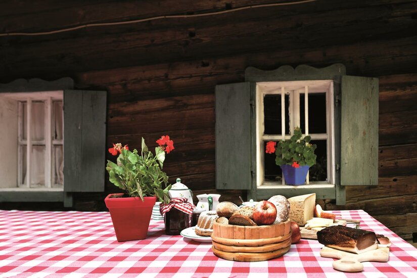 <p>Ein Tisch mit rot-weißen Tischdecken, auf dem verschiedene Lebensmittel und eine Blume stehen. Im Hintergrund sieht man Holzwände und Fenster mit grünen Fensterläden.</p>