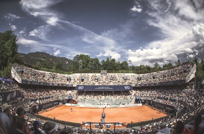 Tennisstadion Ein Tennisplatz mit roten Sand und vielen Zuschauern auf den Tribünen. Der Himmel ist klar und es sind einige Wolken zu sehen.