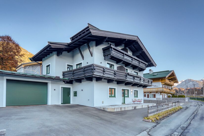 Modernes alpines Wohnhaus mit weißer Fassade, dunklen Holzbalkonen und einer Garage an einer asphaltierten Straße.