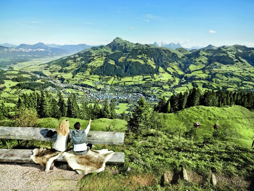 Aussichtspunkt am Hahnenkamm Zwei Personen sitzen auf einer Holzbank und blicken auf ein Tal mit Bergen im Hintergrund.