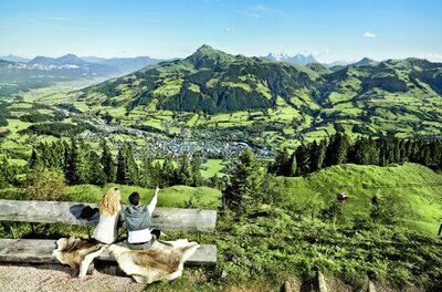 Zwei Personen sitzen auf einer Holzbank und blicken auf ein Tal mit Bergen im Hintergrund.