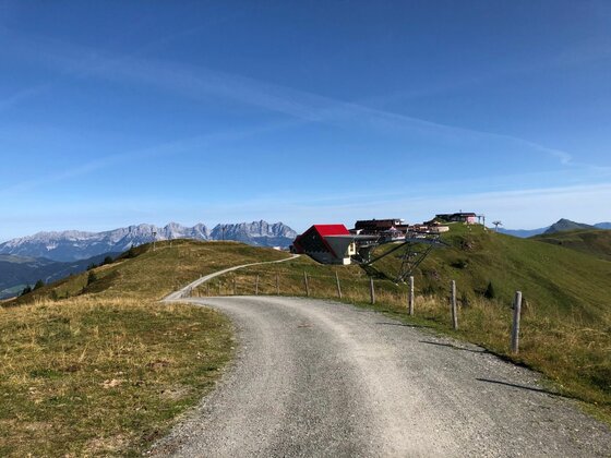 KAT Walk Etappe 4 Schotterweg führt über eine Almwiese zu einer Bergbahnstation und Berghütte vor Gebirgskulisse.