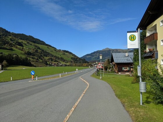 Bushaltestelle an einer Landstraße vor einer Bergkulisse mit grünen Wiesen und blauem Himmel.