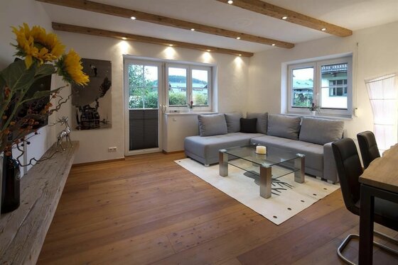 Wohnbereich Appartement Ski Rent Michael Modernes Wohnzimmer mit grauem Ecksofa, Glastisch und Holzboden unter einer Decke mit Holzbalken.
