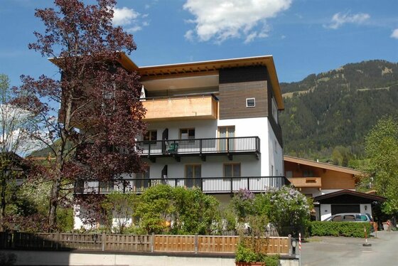 Mehrstöckiges Gästehaus mit Holzbalkonen vor bewaldetem Berghang unter blauem Himmel.