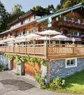 Mehrstöckiger Gasthof aus dunklem Holz mit großer Sonnenterrasse und weißen Schirmen.