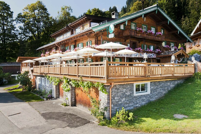 Mehrstöckiger Gasthof aus dunklem Holz mit großer Sonnenterrasse und weißen Schirmen.