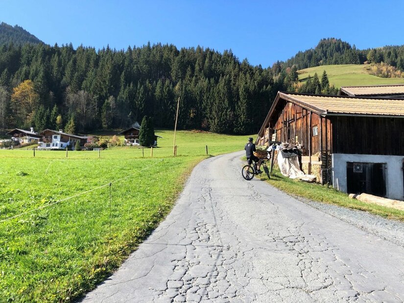 Ein Mountainbiker auf einer asphaltierten Straße neben einem Holzstadel vor bewaldeten Bergen.