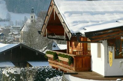 Verschneites Holzhaus mit Balkon vor einer Dorfkirche im Winter.