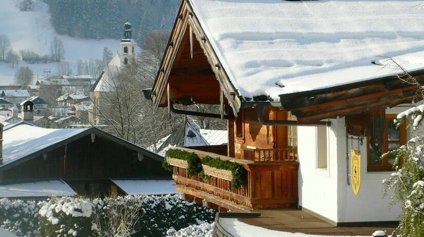 Verschneites Holzhaus mit Balkon vor einer Dorfkirche im Winter.