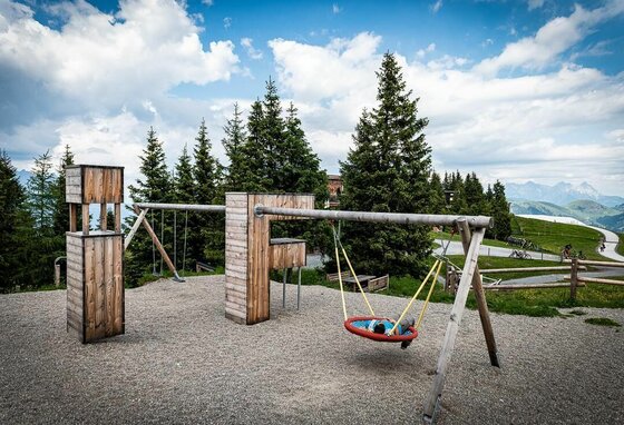 Spielplatz am Berghaus Tirol Ein Kind liegt in einer Nestschaukel auf einem Holzspielplatz vor Nadelbäumen und Bergen.