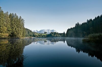 Ein ruhiger See umgeben von Nadelwald mit Spiegelung des Wilden Kaisers im Hintergrund.