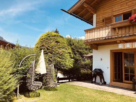 Haus Hagleitner Außenansicht Alpines Holzhaus mit Garten, zwei Hängesesseln auf einer Wiese und einem Grill auf der Terrasse.