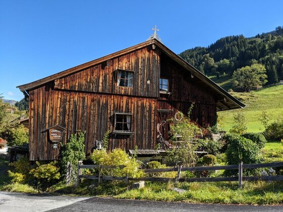 Rustikales Holzhaus mit dunkler Fassade vor bewaldetem Berghang unter blauem Himmel.
