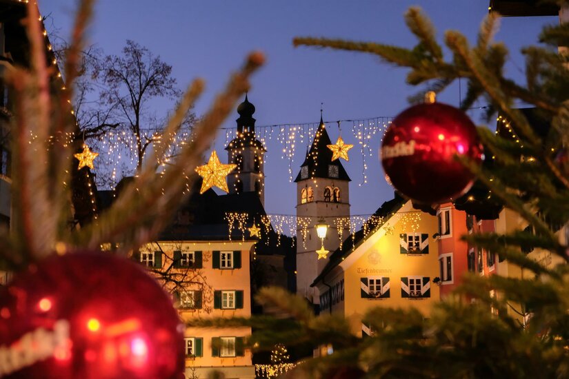 Beleuchtete Kitzbüheler Altstadt bei Dämmerung mit Kirchtürmen, Lichterketten und roten Christbaumkugeln im Vordergrund.