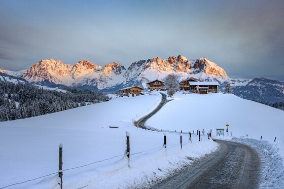 Verschneite Berglandschaft mit einem Bauernhof und einer kurvigen Straße vor einem Gebirgsmassiv.