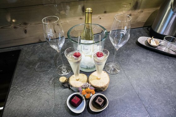 Sektflasche im Kühler, zwei gefüllte Sektgläser mit Himbeeren und Pralinen auf einer dunklen Arbeitsplatte.