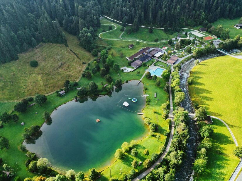 Eine malerische Landschaft mit einem See, umgeben von grünen Wiesen und Bäumen. Im Hintergrund sind Gebäude und ein Schwimmbad sichtbar.