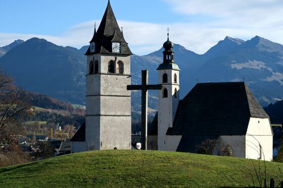 Weiße Kirche mit zwei Türmen und großem Holzkreuz vor einer Gebirgskette.