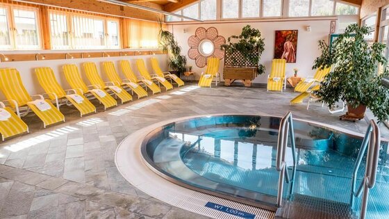 Wellness Center Ein entspannender Wellnessbereich mit einem runden Whirlpool und gelben Liegen. Große Fenster lassen viel Tageslicht herein und schaffen eine ruhige Atmosphäre.