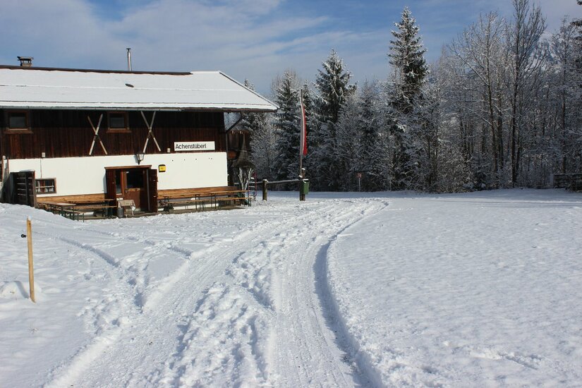 Ein verschneites Holzhaus mit weißer Fassade und schneebedecktem Dach neben einer präparierten Winterspur.