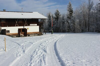 Ein verschneites Holzhaus mit weißer Fassade und schneebedecktem Dach neben einer präparierten Winterspur.