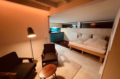 Moderner Wohn- und Schlafbereich mit zwei Sesseln, Holztisch und Doppelbett vor einer Küchenzeile.
