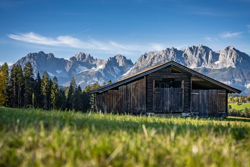 Hochzeit - Huestadl <p>Eine rustikale Holzhütte steht vor majestätischen Bergen, dem Wilden Kaiser. Die Landschaft ist grün und einladend mit strahlendem Himmel.</p>